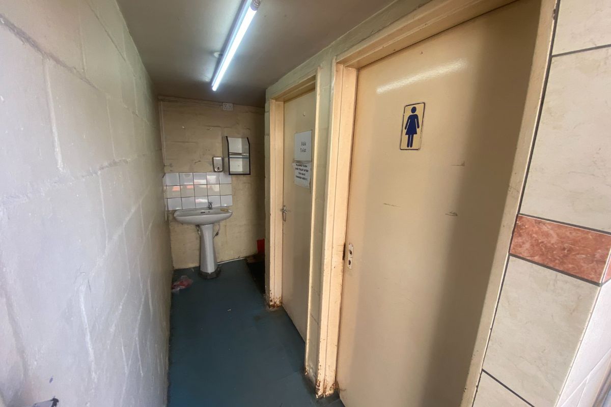 Industrial restroom facility featuring a pedestal basin, tiled splashback, and separate toilet cubicles with basic fluorescent lighting.