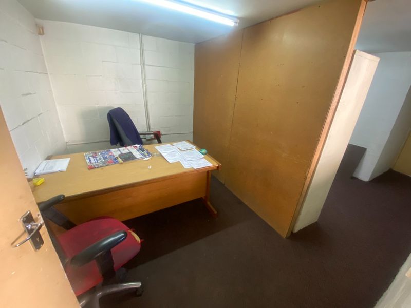A small industrial office space featuring painted concrete block walls, fluorescent lighting, and carpet flooring, suitable for a private workspace.