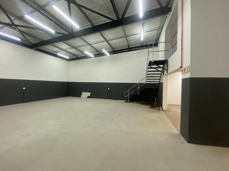Spacious industrial warehouse unit featuring high ceilings with exposed steel trusses, concrete flooring, and a metal staircase leading to an upper level.