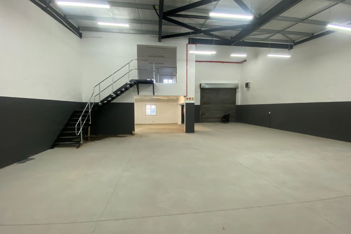 Spacious industrial warehouse unit featuring high ceilings with exposed steel trusses, concrete flooring, a roller shutter door, and an internal steel staircase.