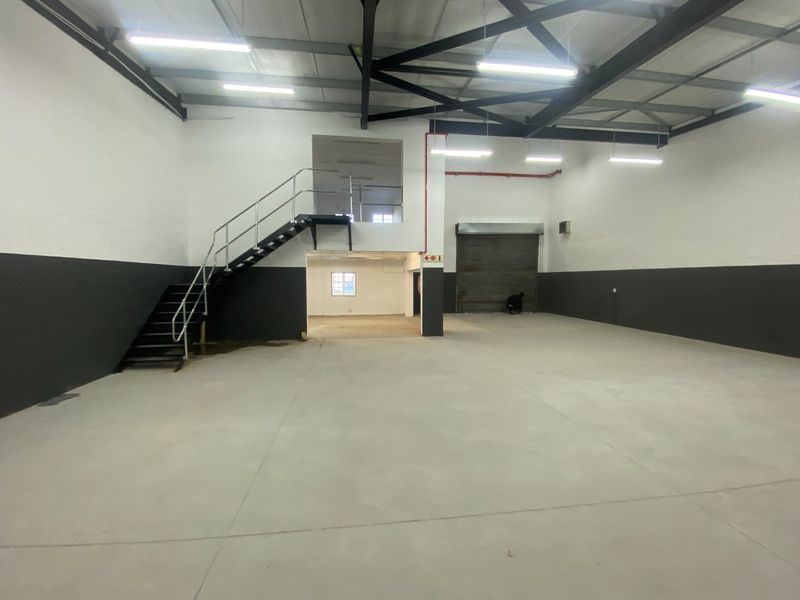 Spacious industrial warehouse unit featuring high ceilings with exposed steel trusses, concrete flooring, a roller shutter door, and an internal steel staircase.