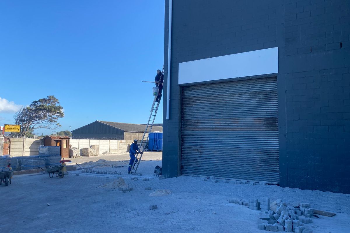 Rear exterior view of an industrial property featuring a large roller shutter door, downpipe, and ongoing paving work in the yard area.