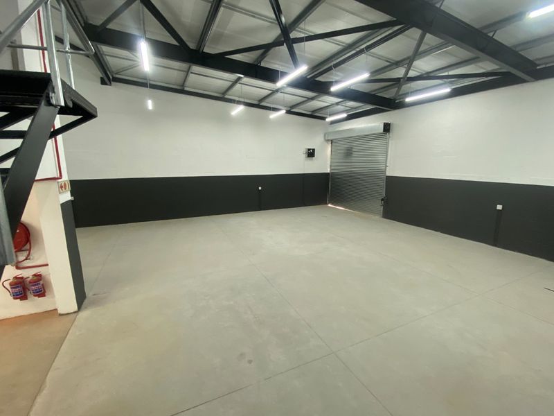 Spacious industrial utility area featuring high ceilings with exposed steel trusses, concrete flooring, and a secure roller shutter door.