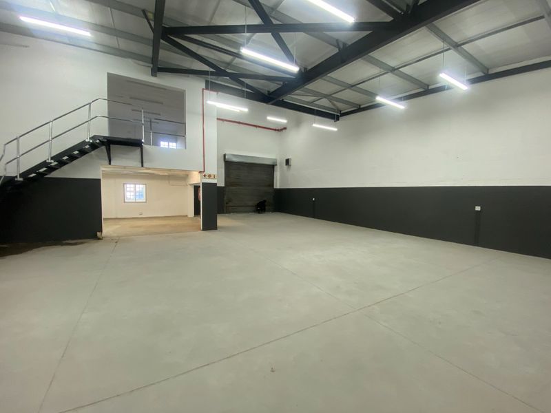 Spacious industrial warehouse unit featuring high ceilings with exposed steel trusses, a mezzanine level with staircase access, and a large roller shutter door.