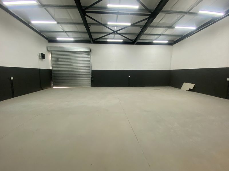 Spacious industrial warehouse unit featuring high ceilings with exposed steel trusses, concrete flooring, and a large roller shutter door for easy access.