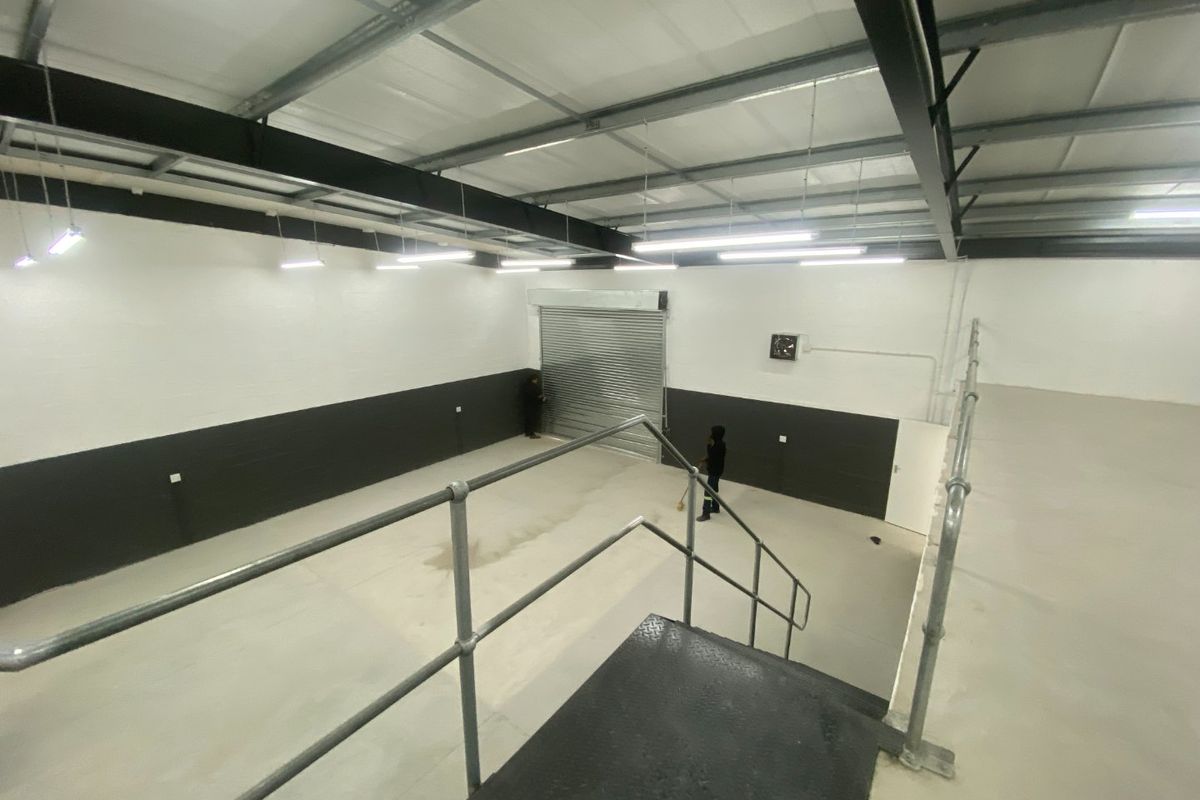 Spacious industrial warehouse unit featuring high ceilings with exposed steel trusses, concrete flooring, a large roller shutter door, and mezzanine access via metal stairs.