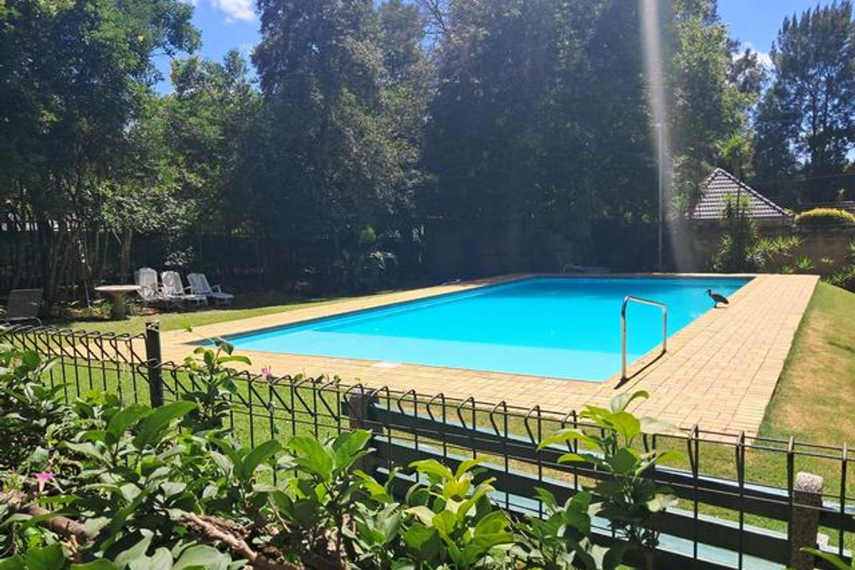 A well-maintained outdoor swimming pool area featuring a paved surround, metal safety fencing, and established garden surroundings.