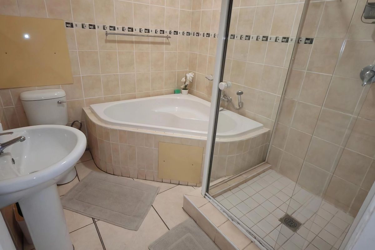 A functional bathroom featuring a corner bathtub, separate shower enclosure, and pedestal basin with neutral wall and floor tiling.