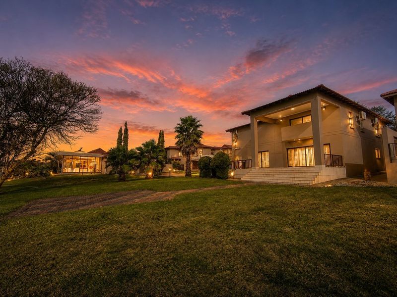 A beautiful rear view of the property at sunset, featuring a spacious lawn, landscaped garden, and a covered patio with an upper-level balcony.
