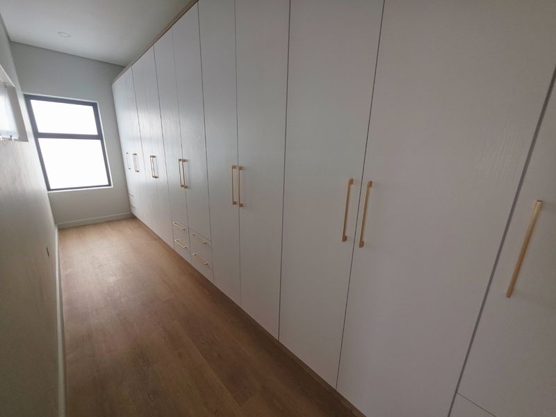 A modern, empty bedroom featuring extensive floor-to-ceiling built-in cupboards with gold-toned handles and light wood-look laminate flooring.