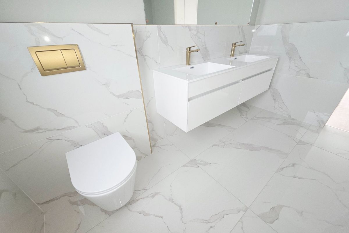 Modern bathroom featuring a wall-mounted double vanity with gold-finish faucets, a wall-hung toilet with matching flush plate, and elegant marble-look porcelain tiling.