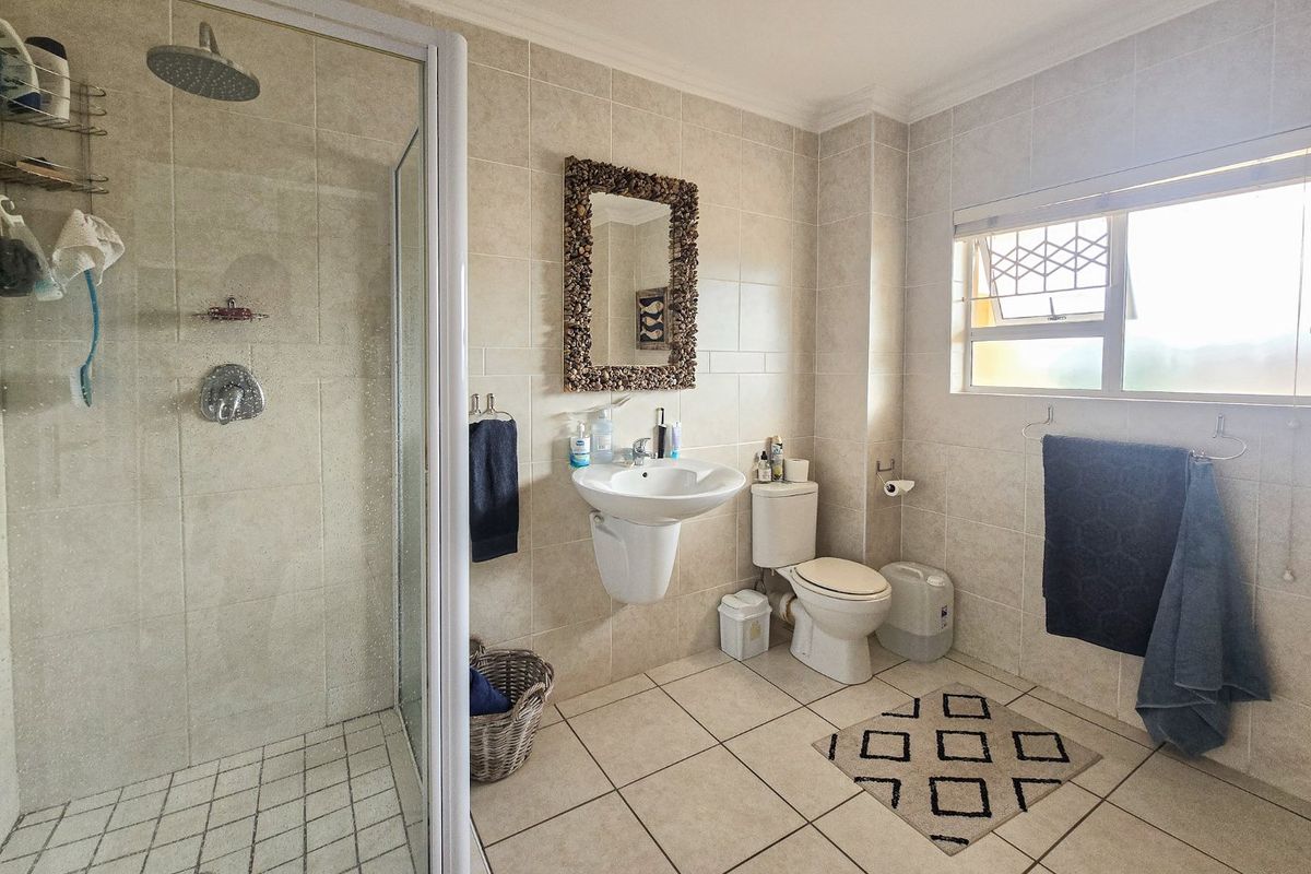 A well-maintained bathroom featuring a glass-enclosed shower, wall-mounted basin, toilet, and neutral floor-to-ceiling tiling with natural light from a side window.