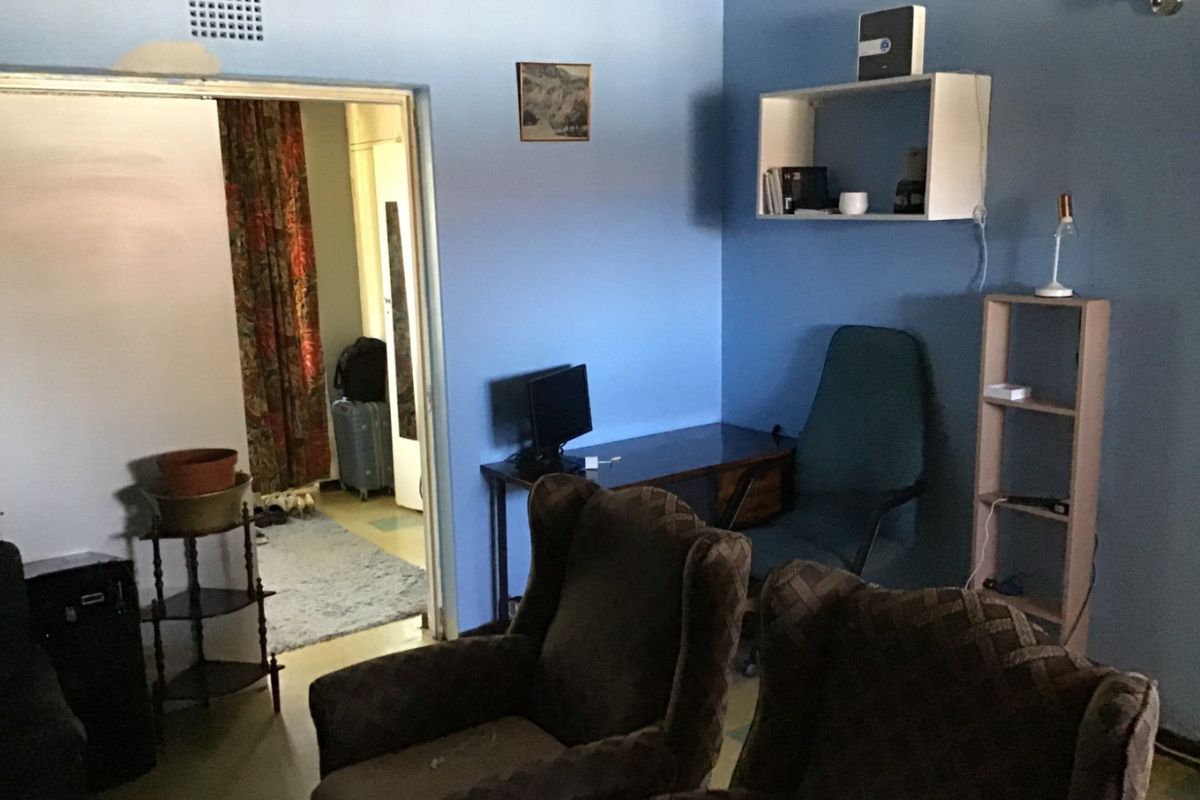 A living area featuring blue walls, a ventilation grille, and built-in wall shelving, showing signs of wear and dated finishes.