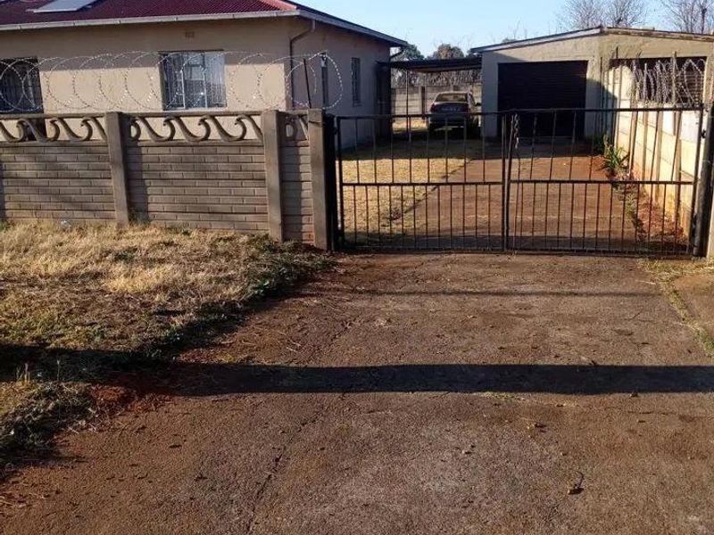 Front view of a single-story house featuring a concrete driveway, metal security gate, perimeter wall with razor wire, and a detached garage.