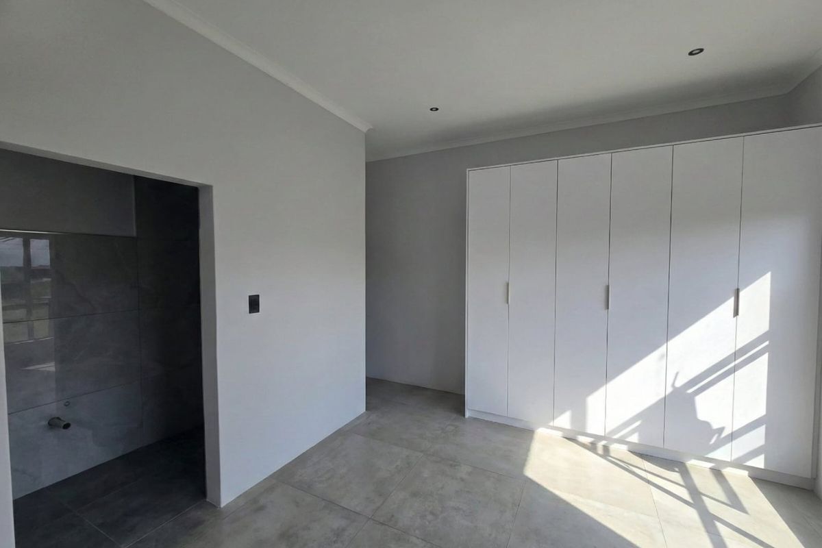A bright, modern bedroom featuring neutral grey floor tiles, built-in white cupboards, and an open-plan en-suite area with contemporary wall tiling.