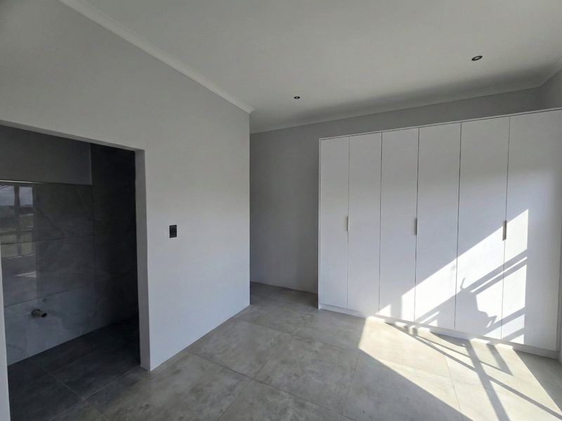 A bright, modern bedroom featuring neutral grey floor tiles, built-in white cupboards, and an open-plan en-suite area with contemporary wall tiling.