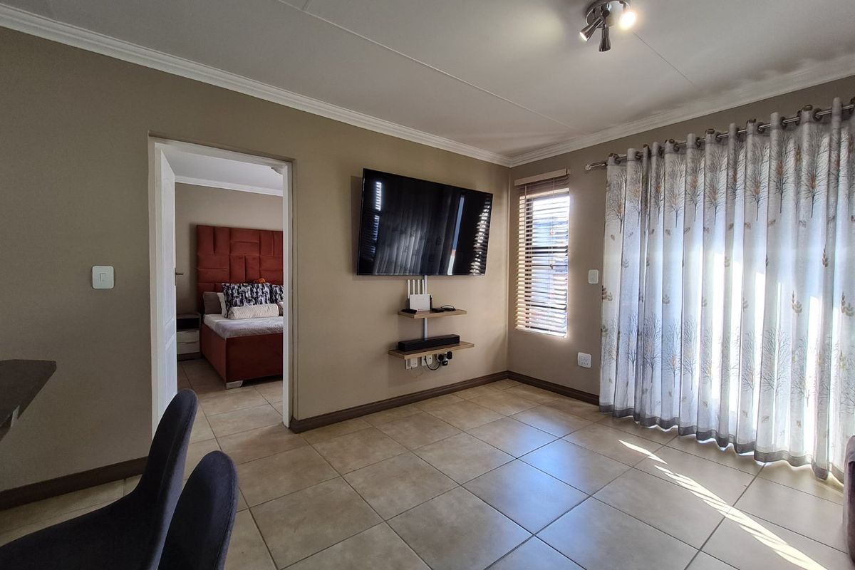 A well-maintained living area featuring neutral tiled flooring, wall-mounted shelving, and large windows with integrated blinds and curtains, leading into an adjacent bedroom.