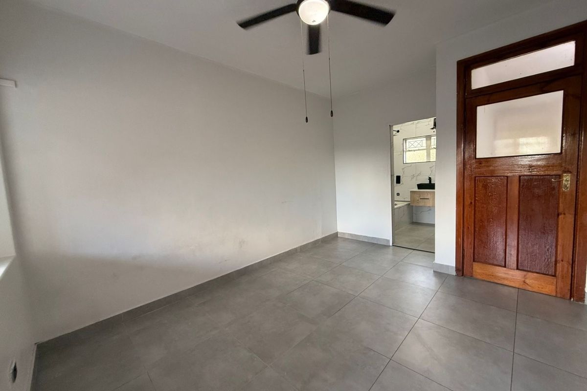 An empty bedroom featuring grey tiled flooring, a ceiling fan, and a traditional wooden door, with an adjoining bathroom visible through the doorway.