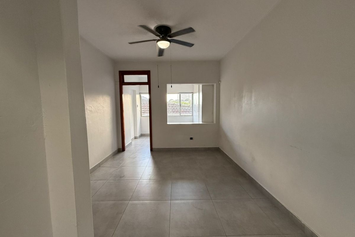 An empty living room featuring neutral tiled flooring, a ceiling fan, and a pass-through window connecting to an adjacent space.