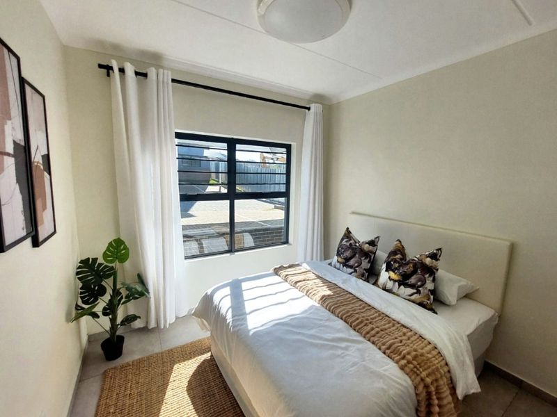 A well-lit bedroom featuring neutral wall tones, tiled flooring, and a large window with security bars, providing a clean and comfortable living space.