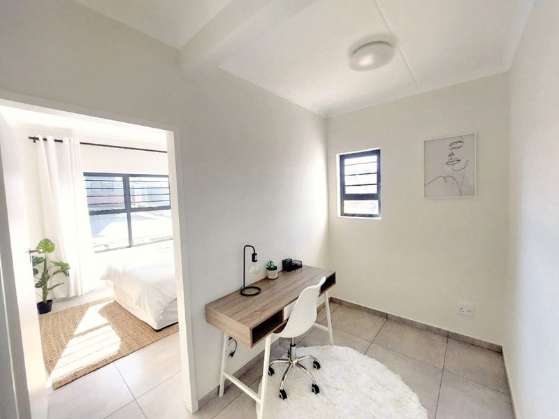 A bright and neutral home office space featuring tiled flooring and natural light, with an open transition to the adjacent bedroom area.