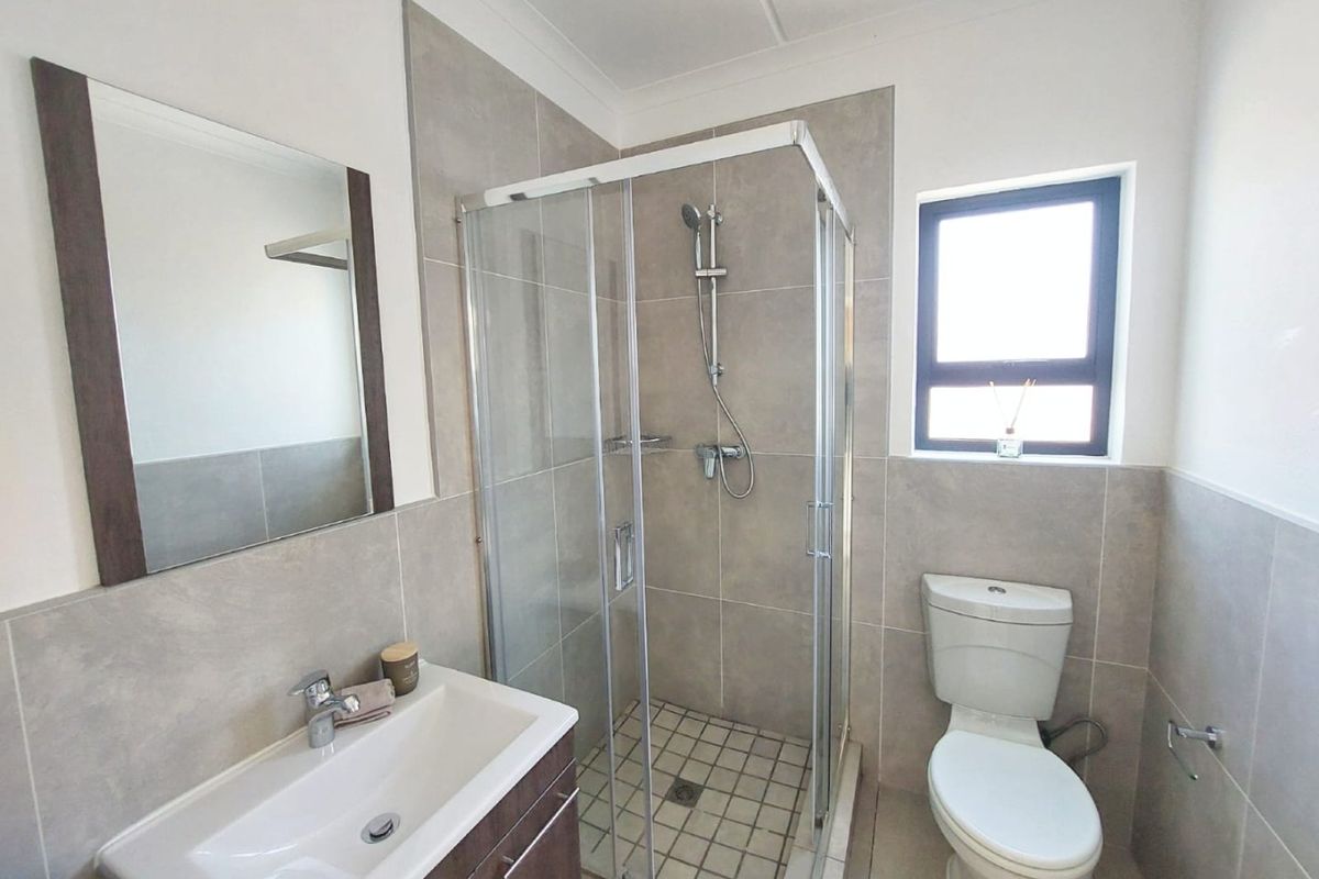 A modern bathroom featuring a glass-enclosed shower, a vanity with a washbasin, a wall-mounted mirror, and a toilet with neutral wall and floor tiling.