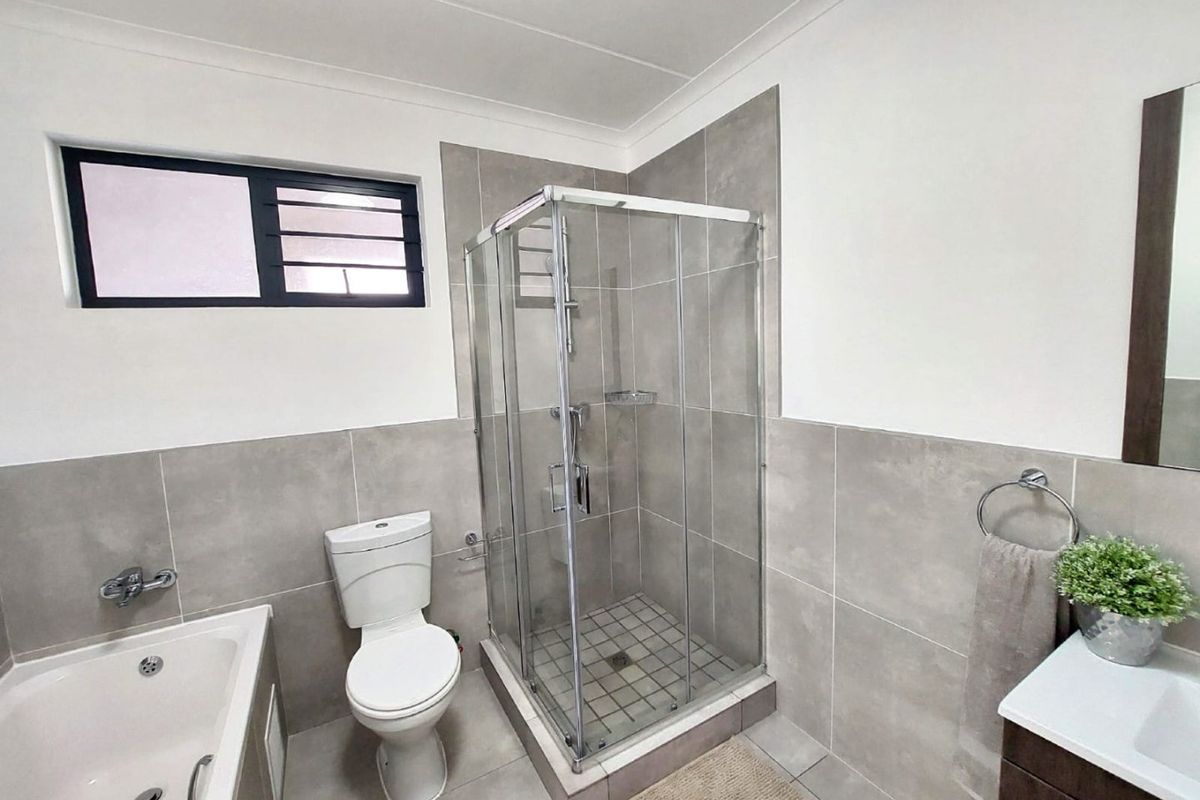 Modern bathroom featuring a glass shower enclosure, bathtub, toilet, and vanity with neutral grey wall and floor tiling.