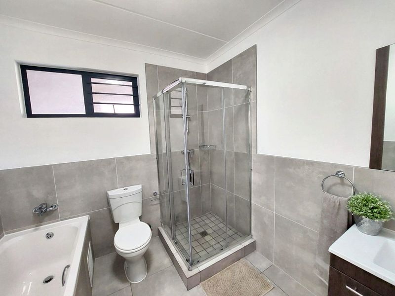 Modern bathroom featuring a glass shower enclosure, bathtub, toilet, and vanity with neutral grey wall and floor tiling.