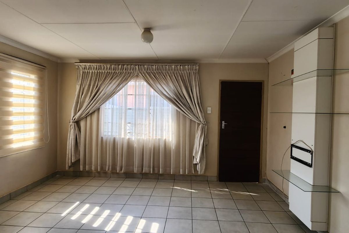 A bright, empty living room featuring tiled flooring, a built-in shelving unit, and large windows with fitted blinds.