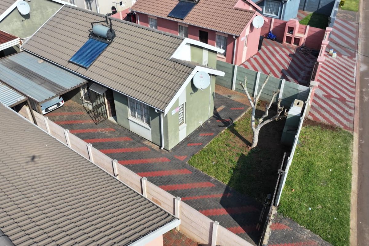 Aerial view of a residential property featuring a tiled roof with solar water heating, a paved driveway, carport, and enclosed garden area.