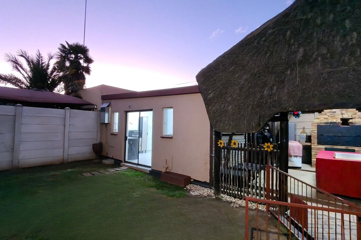 Rear garden area featuring a thatched lapa with a built-in braai, paved patio, and access to the main dwelling via a sliding glass door.