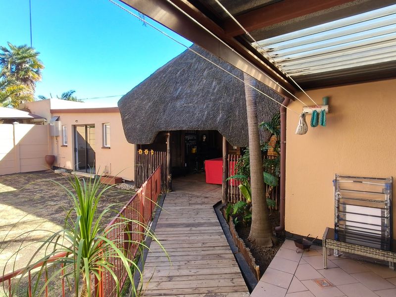 A rear exterior view featuring a thatched roof structure, wooden walkway, tiled patio area, and a small garden space.