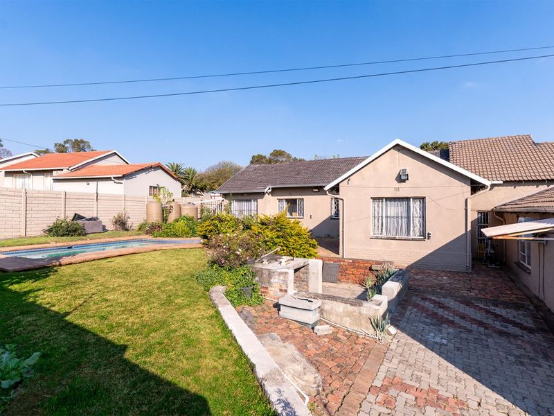 Rear exterior view featuring a swimming pool, brick-paved patio area with a built-in braai, and a landscaped garden with boundary walling.