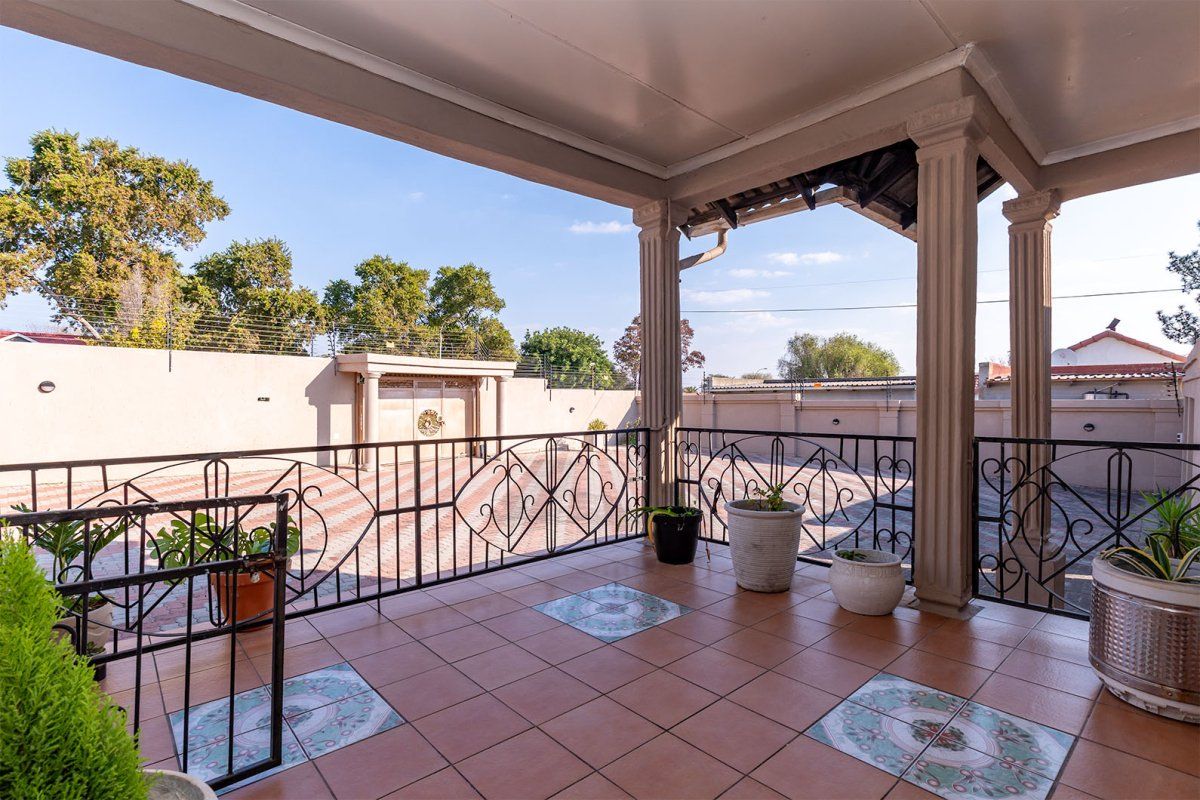 A covered patio area featuring tiled flooring, decorative columns, and a wrought iron balustrade, overlooking a paved driveway and perimeter wall.
