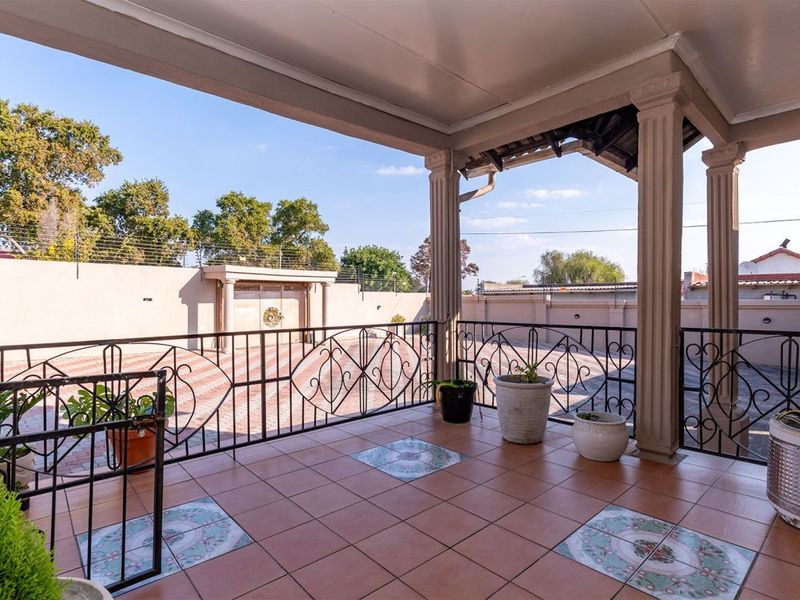 A covered patio area featuring tiled flooring, decorative columns, and a wrought iron balustrade, overlooking a paved driveway and perimeter wall.