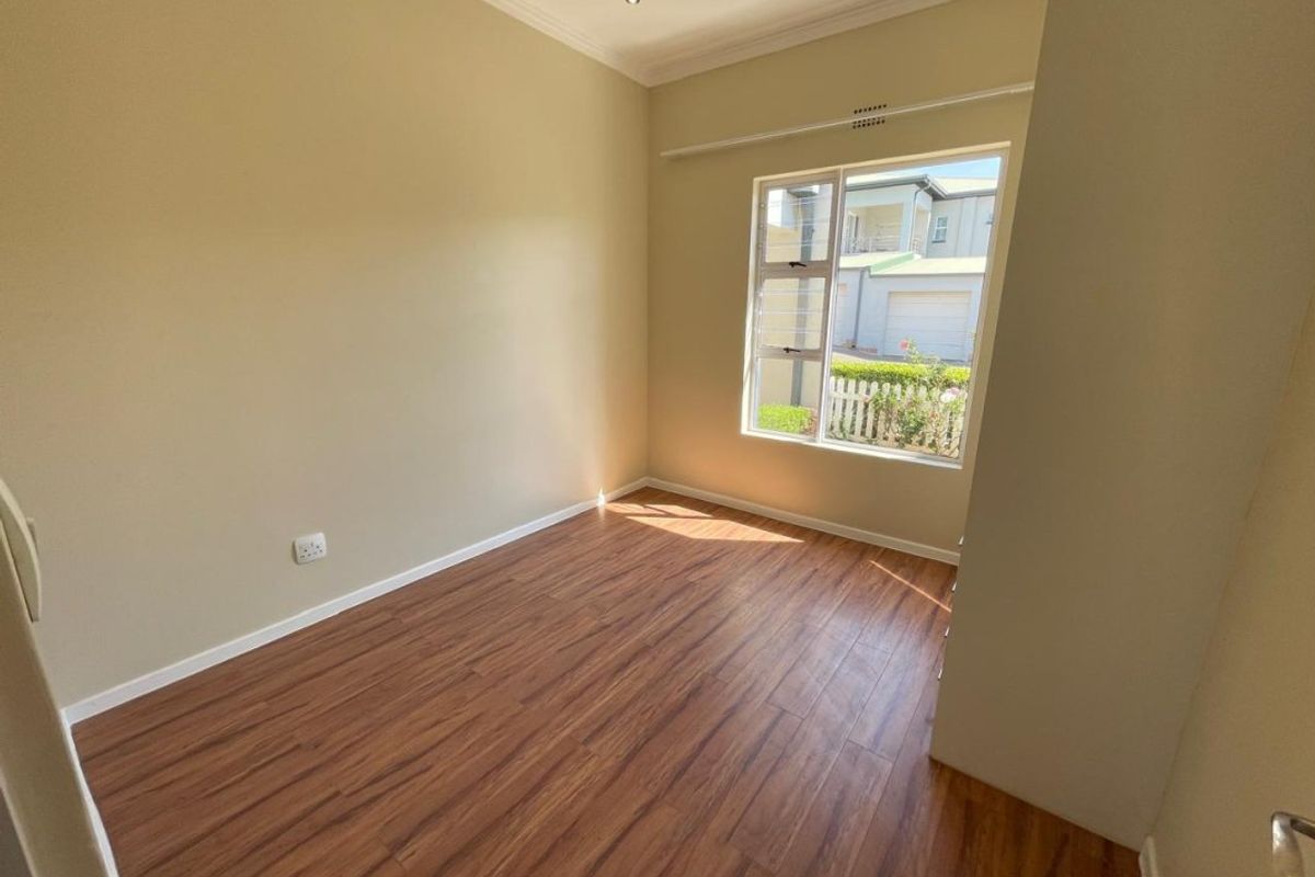 A bright, empty bedroom featuring modern laminate flooring, neutral wall tones, and a large window providing natural light and a view of the exterior.