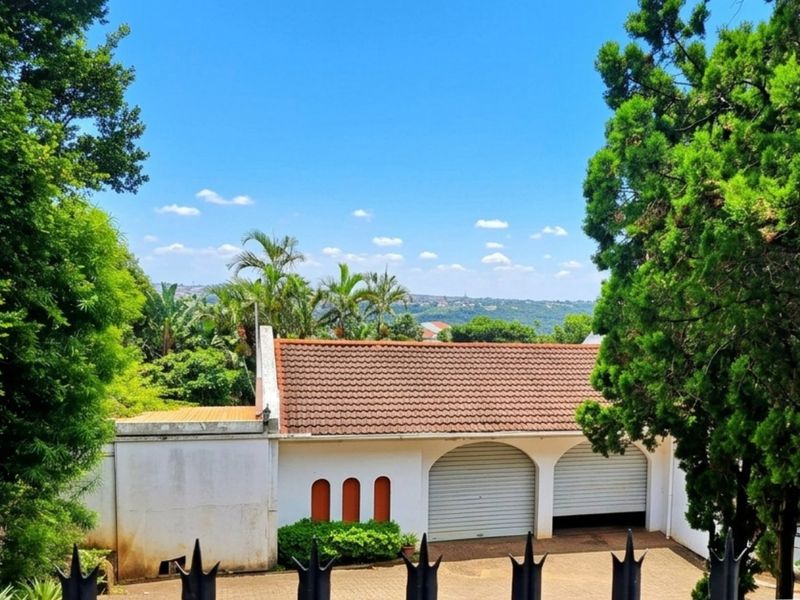 Front view of the property featuring a double garage with arched openings, a tiled roof, and a paved driveway surrounded by mature trees.