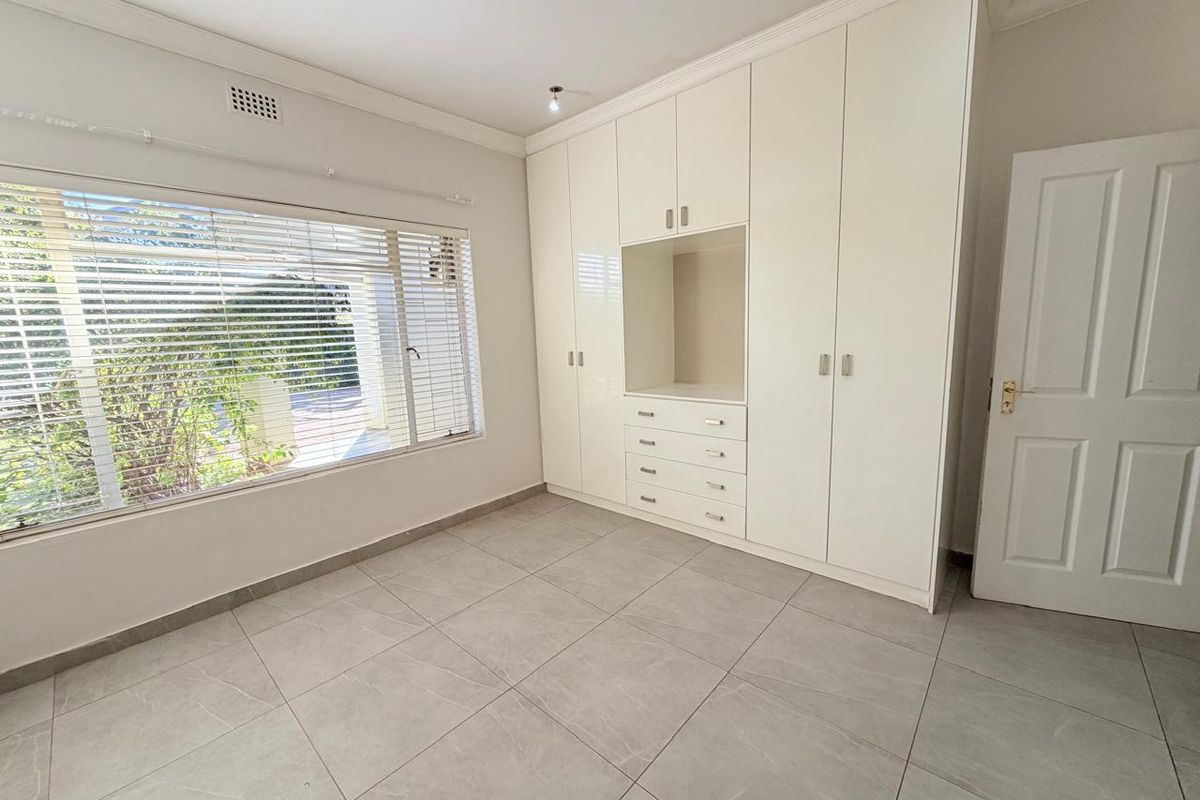 A bright, empty bedroom featuring neutral tiled flooring, built-in floor-to-ceiling cupboards, and large windows with fitted venetian blinds.