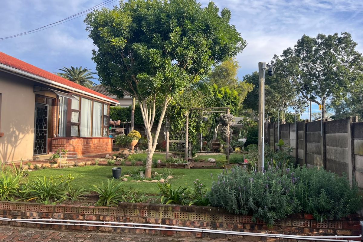 A rear garden view featuring a lawn, mature trees, a brick boundary wall, and a paved area adjacent to the house structure.