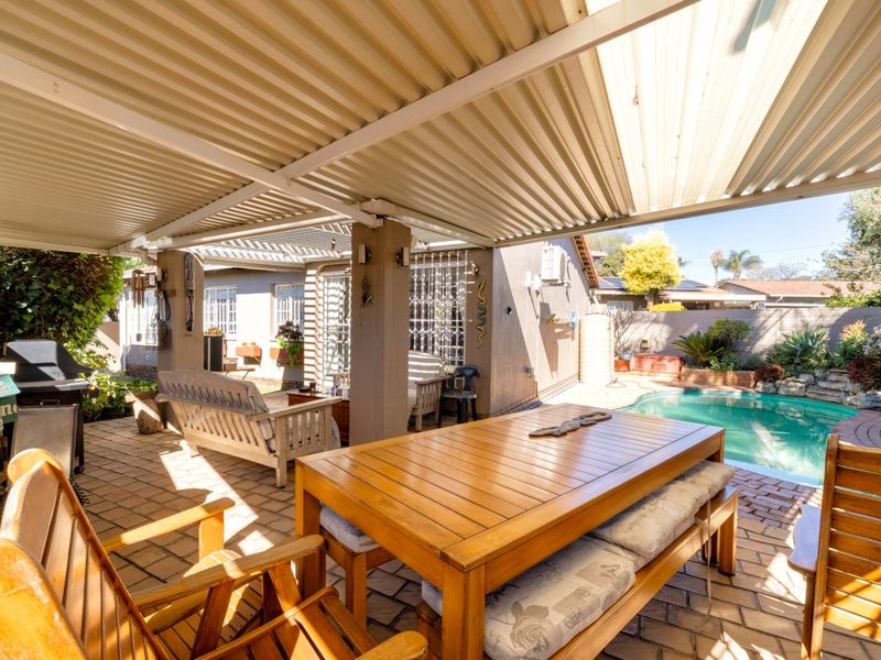 Covered outdoor patio area featuring brick paving and a corrugated metal roof, overlooking a private swimming pool and garden space.