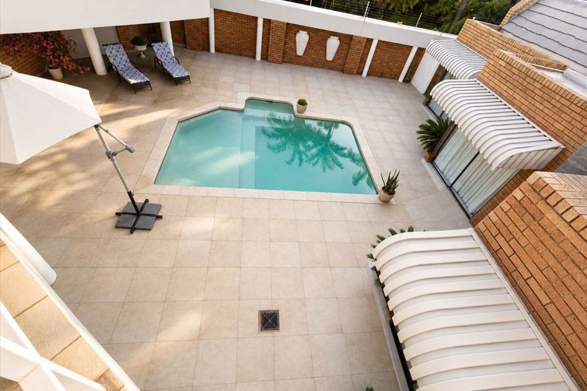 An elevated view of a paved courtyard featuring a swimming pool, brick boundary walls, and white retractable awnings over the building access points.