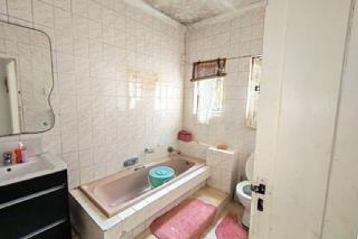 A dated bathroom featuring a built-in bathtub, wall-mounted basin, and toilet with fully tiled walls and floor, requiring cosmetic updates.