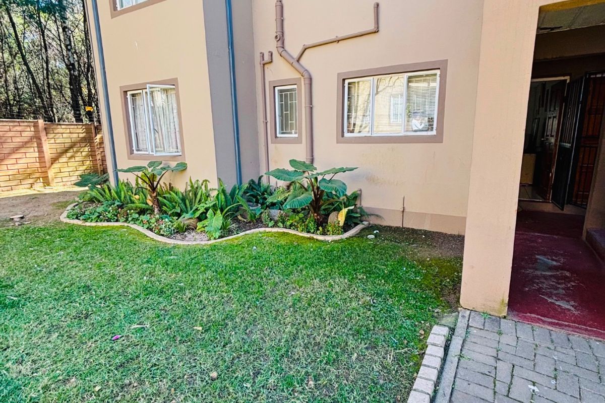 Rear exterior view of the apartment building featuring a small garden bed, paved walkway, and visible external plumbing infrastructure.