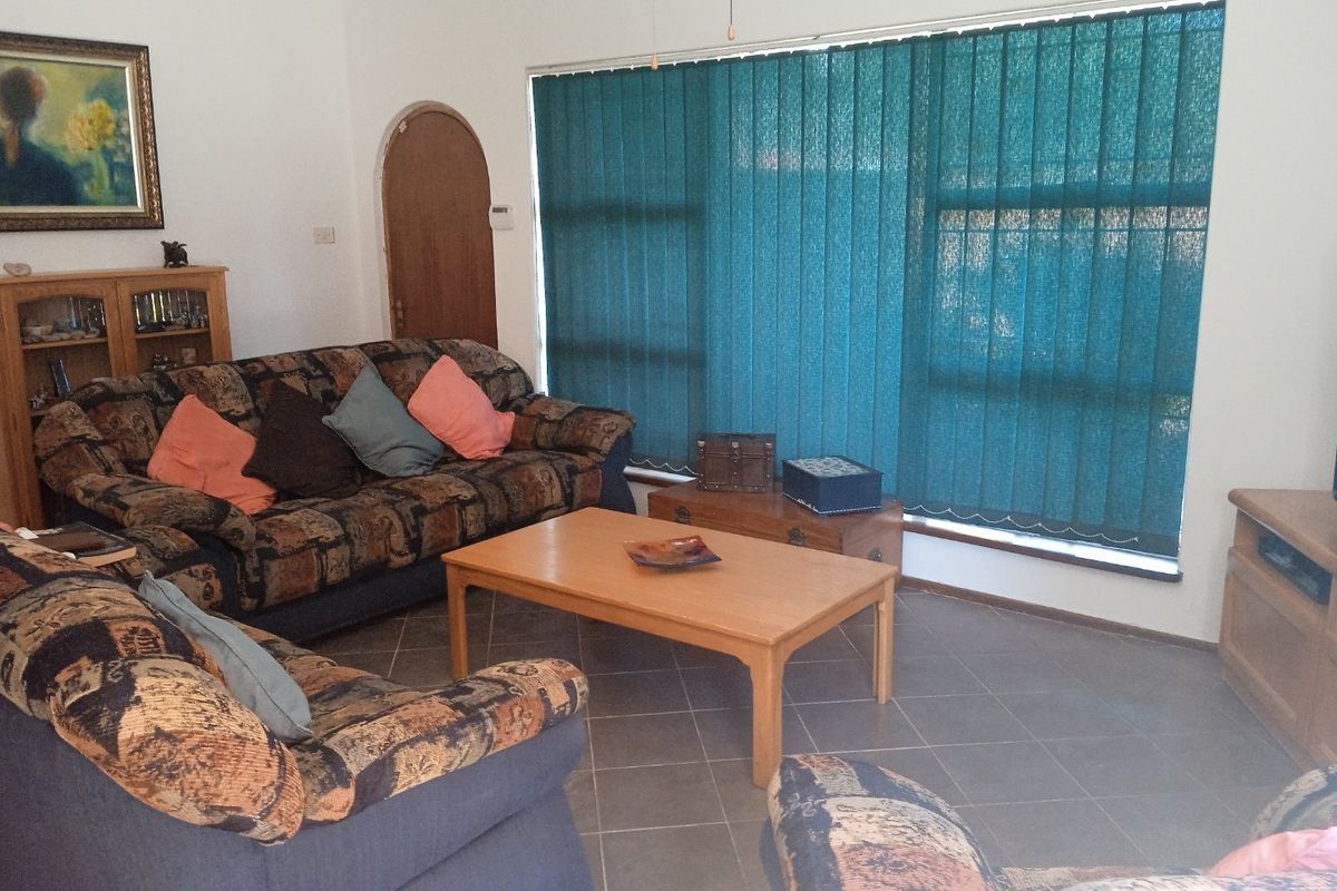 A living room featuring tiled flooring, an arched doorway, and large windows fitted with vertical blinds.