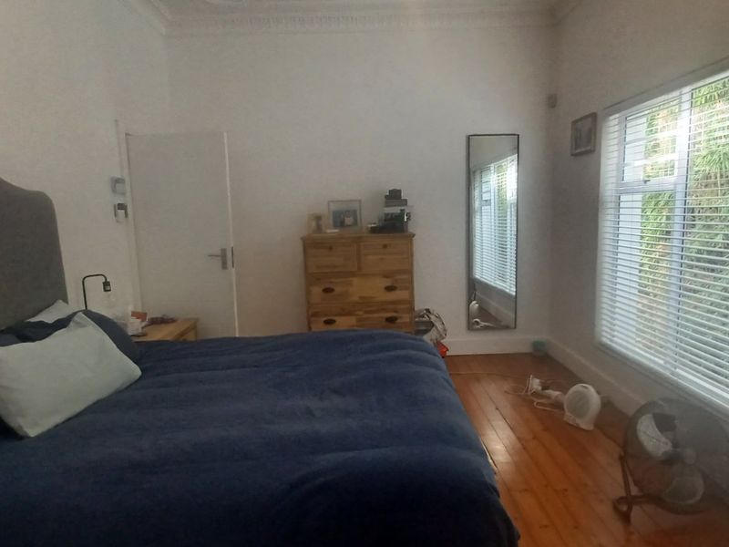 A spacious bedroom featuring polished timber flooring, decorative cornice detailing, and large windows fitted with venetian blinds overlooking greenery.