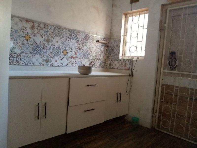 A functional kitchen area featuring white built-in cabinetry, a decorative patterned tile backsplash, and a security gate, currently requiring cosmetic wall repairs.