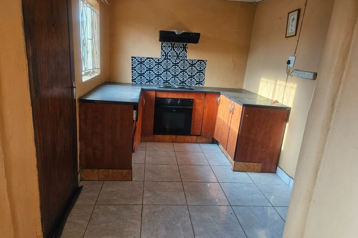 A compact kitchen featuring built-in wooden cabinetry, dark countertops, integrated oven and hob, and tiled flooring, offering a functional space ready for modernization.