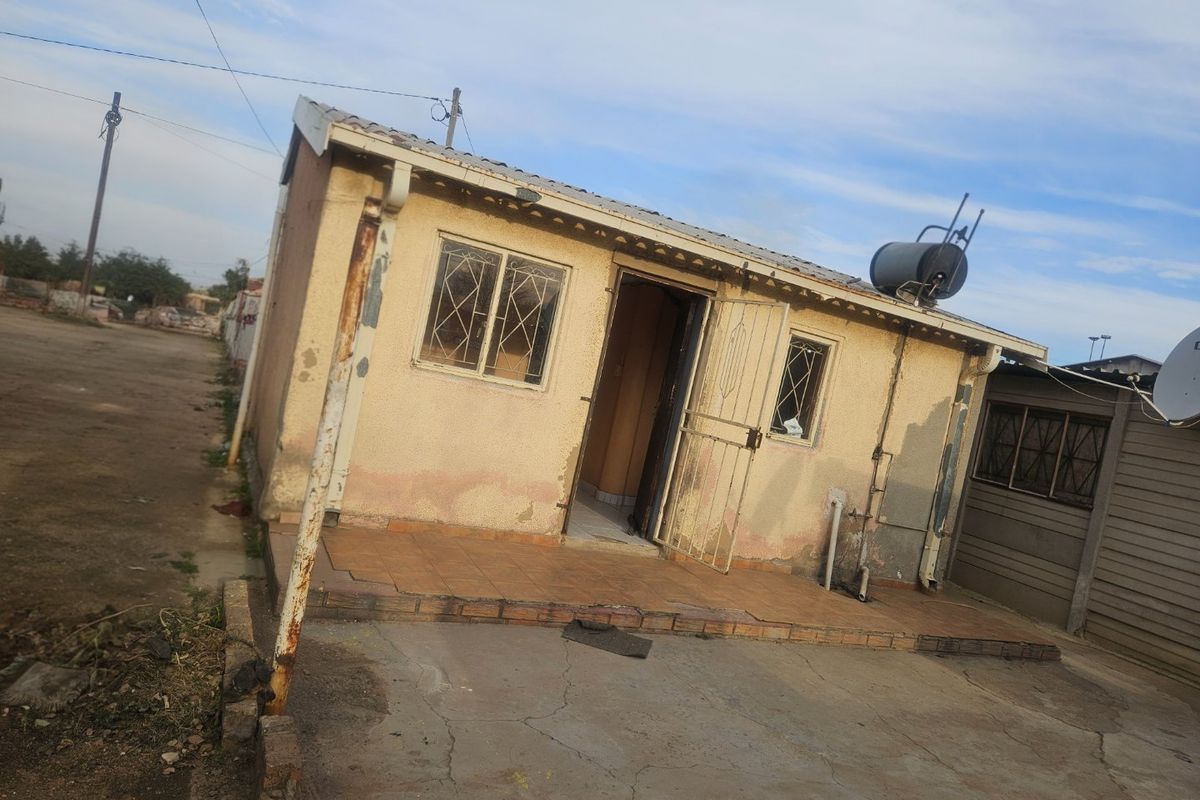 Exterior view of a small residential dwelling featuring a tiled patio, security gates, and a solar water heater, showing signs of significant exterior wear.
