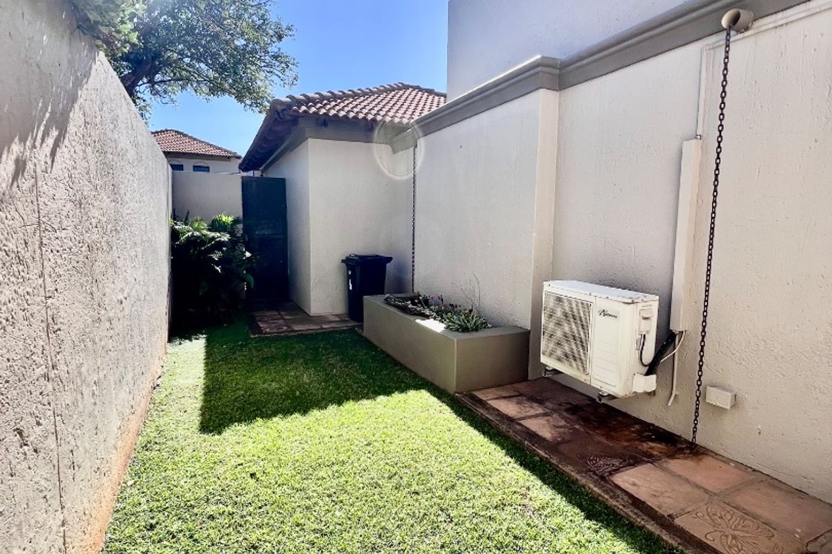 A private rear courtyard featuring a small lawn, raised garden bed, paved walkway, and wall-mounted air conditioning unit.
