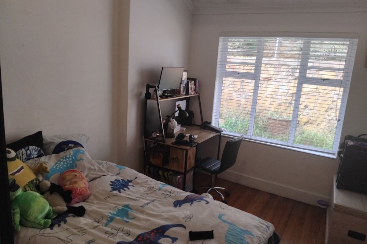 A bedroom featuring timber flooring, cove cornice detailing, and a large window fitted with venetian blinds overlooking an exterior stone retaining wall.